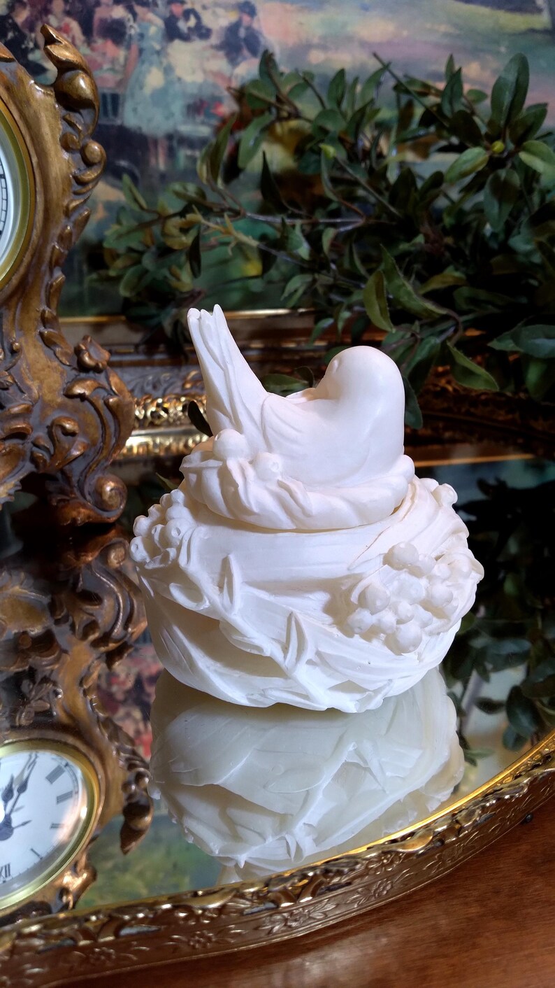 May include: A white ceramic bird figurine perched on a nest-shaped base. The bird has a smooth, rounded body and is nestled among detailed floral and leaf accents. The figurine is displayed on a reflective surface, with a gold-framed clock in the background.