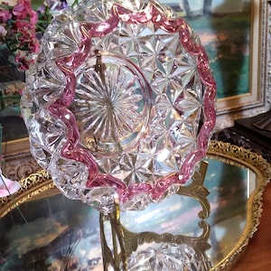 May include: A clear glass dish with a pink rim, featuring a cut-glass design. The dish is displayed on a gold-colored stand.