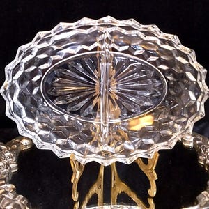 May include: A clear, divided glass serving dish with a diamond-cut pattern. The oval-shaped dish sits on a gold-colored metal stand with ornate detailing. The dish is designed to hold two separate items.