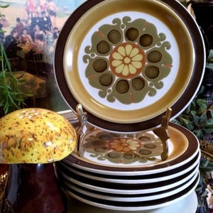 May include: A stack of brown and white ceramic plates with a floral design. The plates have a brown rim and a white center with a green, brown, and yellow floral pattern. The plates are stacked on a gold-colored stand.
