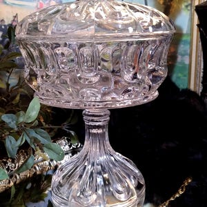 May include: A clear glass candy dish with a lid and a pedestal base. The dish has a decorative pattern of raised circles and the base has a fluted design.