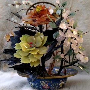 May include: A decorative floral arrangement featuring carved stone flowers in shades of orange, yellow, and pale pink, accented by dark leaves and thin branches, set in a round blue cloisonné bowl with a brass rim and colorful floral patterns.