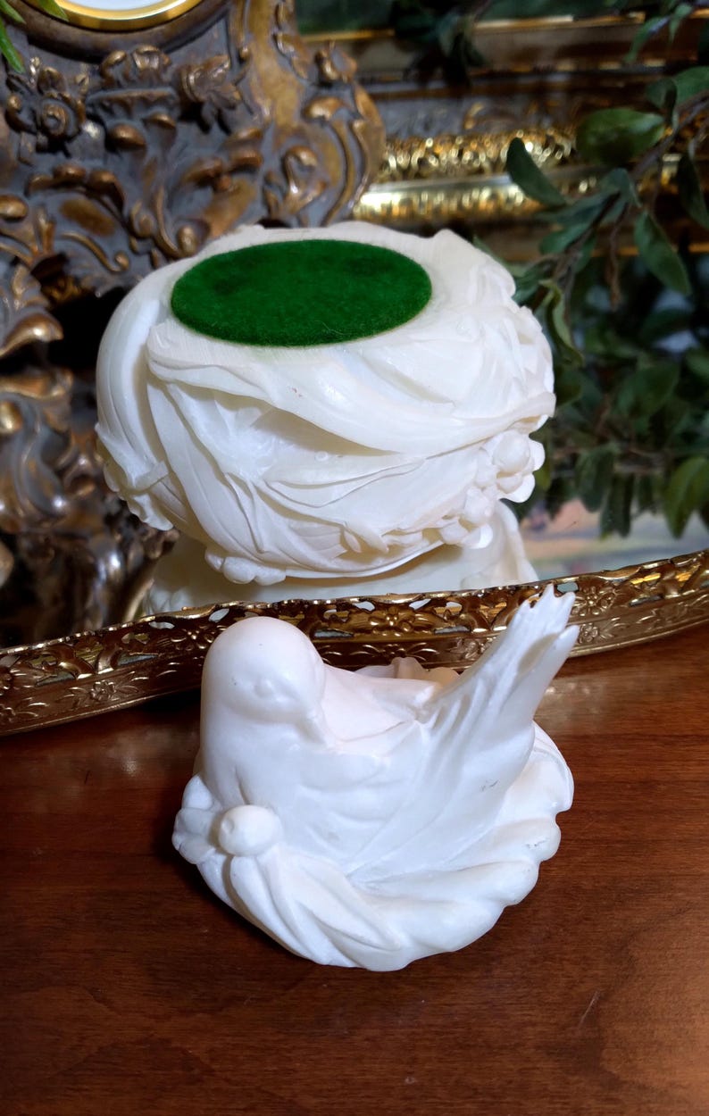 May include: A white, carved sculpture of a bird with outstretched wings, resting on a nest-like base. A separate, matching sculpture with a green, circular insert sits behind it. The sculptures are likely decorative items.