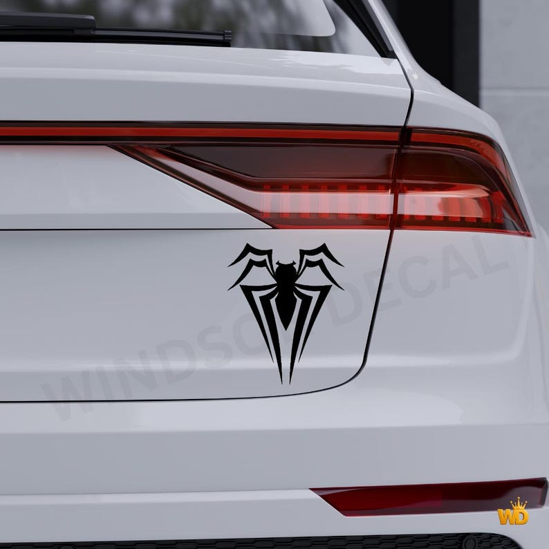 May include: A black spider decal on the back of a white car. The decal features a stylized spider design with sharp, angular lines. The car's taillights are visible above the decal.