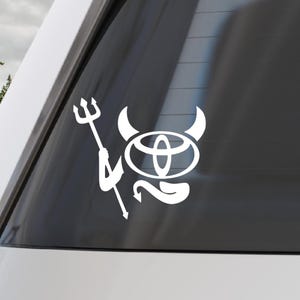 Könnte beinhalten: Weißes Vinyl-Aufkleber mit einem teuflischen Design. Das Design beinhaltet das Toyota-Logo mit Hörnern, einem Schwanz und einem Dreizack. Der Aufkleber ist auf einer Autoscheibe angebracht und zeigt eine verspielte und einzigartige Ästhetik.