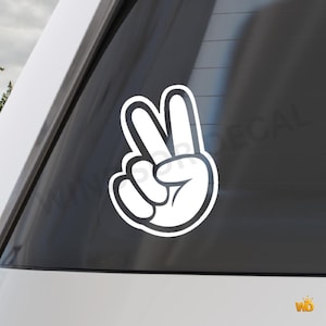 May include: White vinyl decal of a hand making the peace sign, with a thick black outline. The decal is applied to a car window. The image is a close-up of the peace sign.