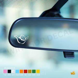 May include: A white vinyl decal featuring a shaka hand gesture graphic applied to a car rearview mirror. Below the mirror, a color selection chart displays options including pink, black, white, ice blue, orange, red, gold, green, and yellow.