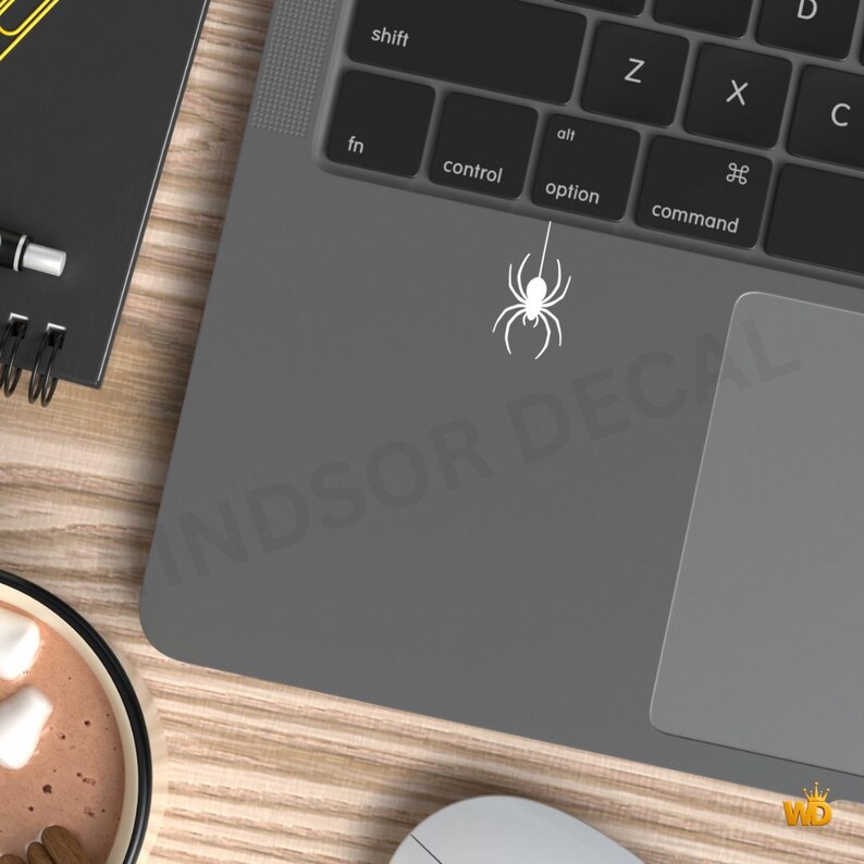 Puede incluir: Un adhesivo de ara&ntilde;a blanco aplicado sobre la superficie gris de un ordenador port&aacute;til, situado justo debajo de la tecla option del teclado. La ara&ntilde;a se representa con una l&iacute;nea vertical que se extiende desde su cuerpo hacia las teclas.