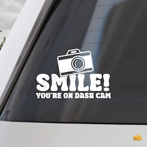 Peut inclure: Autocollant en vinyle blanc avec une illustration d'appareil photo et le texte "SMILE! YOU'RE ON DASH CAM." L'autocollant est conçu pour être appliqué sur une vitre de voiture. Le graphisme de l'appareil photo est un design simple et souligné.