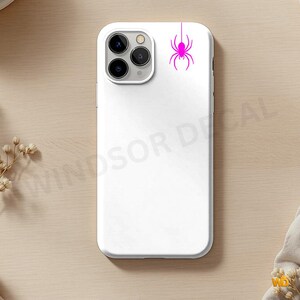 Puede incluir: Una funda de smartphone blanca con un gr&aacute;fico de ara&ntilde;a rosa brillante que cuelga de una l&iacute;nea vertical en la esquina superior derecha. La funda tiene un recorte para un sistema de triple c&aacute;mara en la parte superior izquierda y un acabado liso.