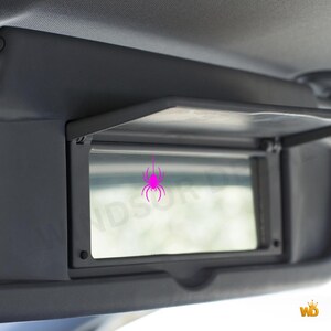 Puede incluir: Un espejo de cortes&iacute;a rectangular integrado en un parasol de coche gris oscuro. La superficie del espejo presenta un peque&ntilde;o gr&aacute;fico de una ara&ntilde;a de color rosa brillante situado en el centro. El parasol est&aacute; abierto, mostrando el cristal.