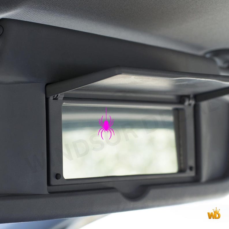 Puede incluir: Un espejo de cortes&iacute;a rectangular integrado en un parasol de coche gris oscuro. La superficie del espejo presenta un peque&ntilde;o gr&aacute;fico de una ara&ntilde;a de color rosa brillante situado en el centro. El parasol est&aacute; abierto, mostrando el cristal.