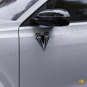 May include: Black spider decal applied to a white car door. The decal features a stylized spider design with a central body and pointed legs. The car's side mirror is visible above the decal.