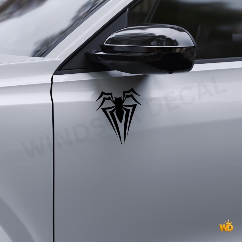 May include: Black spider decal applied to a white car door. The decal features a stylized spider design with a central body and pointed legs. The car's side mirror is visible above the decal.