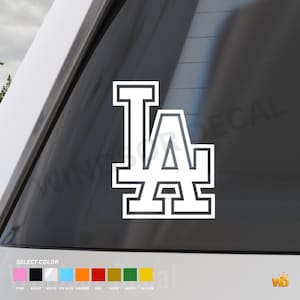 May include: A white vinyl decal featuring the letters LA in a block-style font with a thick outline. The decal is applied to a dark glass surface. Below the decal, a colour selection guide displays small squares in pink, black, white, blue, orange, red, gold, green, and yellow.