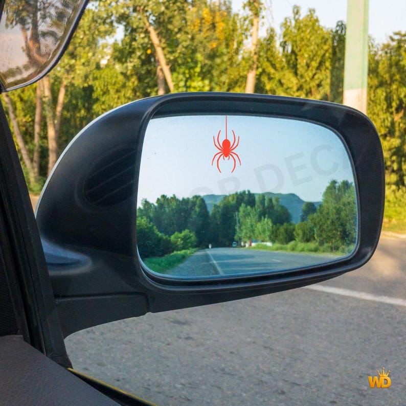 Puede incluir: Un espejo lateral de coche negro con una pegatina de ara&ntilde;a roja aplicada sobre la superficie de cristal. El espejo refleja una carretera rodeada de &aacute;rboles verdes y una cordillera lejana bajo un cielo despejado, con la ara&ntilde;a roja en el centro.