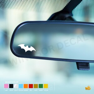 Puede incluir: Una calcomanía de vinilo blanca con la silueta icónica de un murciélago aplicada en un espejo retrovisor de coche. Debajo del espejo, una tabla de selección de colores muestra opciones como rosa, negro, blanco, azul hielo, naranja, rojo, oro, verde y amarillo.