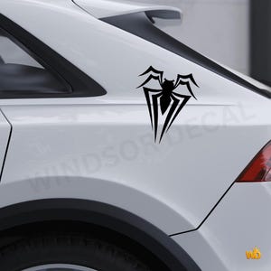 May include: Black spider emblem car decal. The decal is a stylized spider design with sharp lines and a symmetrical shape. The decal is applied to the white body of a car. The text "WINDSOR DECAL" is visible in the background.
