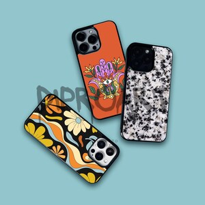 May include: Three different iPhone cases with unique designs. The first case is orange with a floral pattern and a purple hand with an eye. The second case is black with a white and gray marble pattern. The third case is black with a colorful abstract pattern.