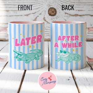 Peut inclure: Deux tasses en céramique avec un motif à rayures verticales bleu clair et blanc et des anses roses. La tasse de gauche présente un alligator vert et le texte rose LATER. La tasse de droite montre un alligator vert avec des lunettes et le texte rose AFTER A WHILE.