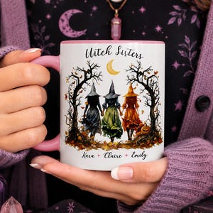 May include: A white ceramic mug with a pink handle and interior, featuring a graphic of three witches in cloaks standing between bare trees under a crescent moon. The text Witch Sisters is printed above, and the names Kara, Claire, and Emily are below.