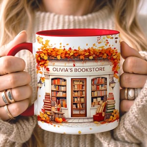 Op de afbeelding: Een witte keramische mok met een rood handvat en binnenkant, voorzien van een illustratie van een boekenwinkel genaamd OLIVIA'S BOOKSTORE, omringd door herfstbladeren, stapels boeken en pompoenen in tinten oranje, geel en bruin.