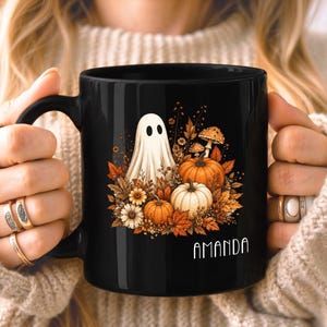 May include: A black ceramic mug featuring a graphic of a white ghost surrounded by orange and white pumpkins, autumn leaves, and mushrooms. The name AMANDA is printed in white capital letters at the bottom right of the graphic design.
