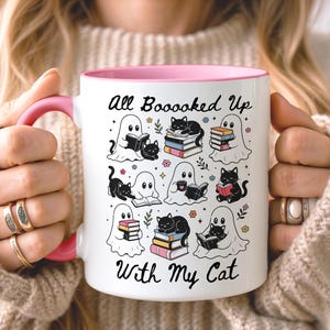 May include: A white ceramic mug with a pink handle and interior rim, featuring a repeating pattern of black cats and white ghosts reading books. The text "All Boooked Up With My Cat" is printed in black script at the top and bottom of the design.