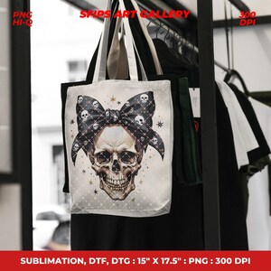 Puede incluir: Un bolso tote blanco con un dise&ntilde;o de calavera y un lazo negro adornado con patrones de calaveras. El bolso, que mide 38 x 44,5 cm, tiene asas negras y se exhibe frente a un perchero. El texto "SFIPS ART GALLERY" es visible.