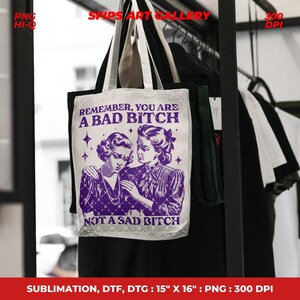 May include: A white tote bag with a purple graphic of two women and the text "REMEMBER, YOU ARE A BAD BITCH NOT A SAD BITCH." The bag is 15" x 16" and has black handles. The bag is hanging on a rack with other clothing.
