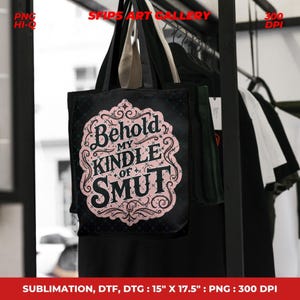 Puede incluir: Bolso tote negro con el texto "Behold My Kindle of Smut" en un dise&ntilde;o ornamental rosa y negro. El bolso mide 38 cm de ancho y 44,5 cm de alto. Cuelga de un perchero con otras prendas.