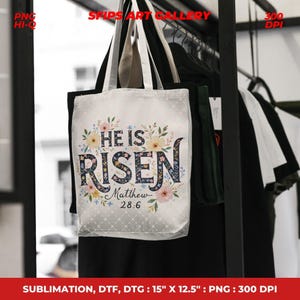 May include: A white tote bag with the text "HE IS RISEN" in a floral design, with "Matthew 28:6" below. The bag is approximately 15 inches by 12.5 inches. The bag is hanging on a black rack with other clothing.