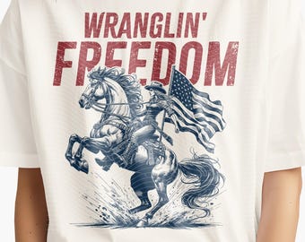May include: White t-shirt featuring a detailed illustration of a woman riding a horse, holding an American flag. The text "WRANGLIN' FREEDOM" is printed above the image in red. The design is suitable for sublimation, DTF, and DTG printing, with dimensions of 15" x 17".