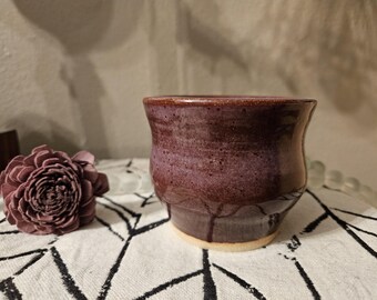 Puede incluir: Una taza de cerámica de color morado oscuro con un esmalte moteado. La taza tiene un fondo redondeado y un labio ligeramente acampanado.