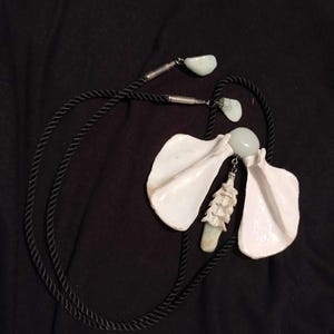 May include: A unique necklace featuring a black rope cord and white, petal-shaped pendants. The necklace includes light green stone accents and a central pendant with a textured, off-white design. The background is a solid black fabric.