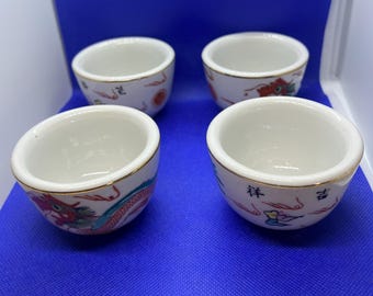 May include: Set of four white ceramic teacups with gold trim and colorful dragon and floral designs. The cups are on a blue surface.