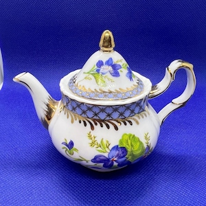 Puede incluir: Una tetera de porcelana blanca con un diseño floral azul y dorado. La tetera tiene una tapa con un pomo dorado y una banda con un patrón azul y dorado alrededor del borde. La tetera está decorada con flores y hojas azules y verdes.