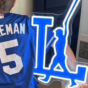 Puede incluir: Una camiseta de béisbol azul con el nombre "FREEMAN" y el número "5" en blanco. Un letrero de neón azul y blanco con la forma del logotipo de los Dodgers de Los Ángeles se sostiene en la mano derecha.