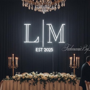 May include: Elegant wedding reception setup with long tables, chairs, and floral arrangements. A neon sign displays the initials "LM" and "EST 2025" against a black backdrop. Crystal chandeliers hang from the ceiling, illuminating the scene.