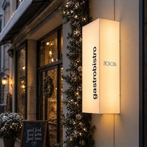 K&ouml;nnte beinhalten: Ein rechteckiges, beleuchtetes Wandleuchtschild mit wei&szlig;er Oberfl&auml;che, das den vertikal ausgerichteten schwarzen Schriftzug "gastrobistro" sowie den kyrillischen Text "ЛЮБОВЬ" in Schwarz zeigt. Das Schild ist an einer hellen Au&szlig;enwand montiert.