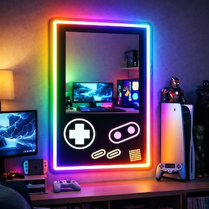 May include: A black mirror in the shape of a retro handheld game console, with a rainbow LED border. The mirror has white controller symbols. The reflection shows a gaming setup. The mirror's dimensions are approximately 76 cm x 46 cm.