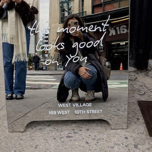 Könnte beinhalten: Ein rechteckiger Standspiegel mit silbernem Rahmen und weißem Schriftzug, der this moment looks good on you, West Village und 169 West 10th Street lautet. Der Spiegel reflektiert eine hockende Person mit Smartphone, die eine dunkle Jacke trägt.