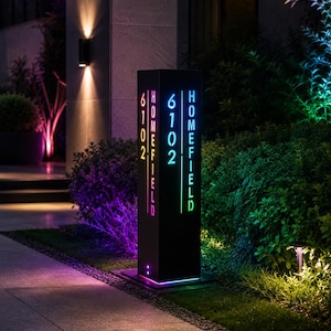 Puede incluir: Un letrero de dirección exterior negro, alto y rectangular, con texto recortado iluminado que dice 6102 HOMEFIELD. El letrero muestra una vibrante iluminación LED multicolor que cambia de azul a verde y rosa, iluminando la base.