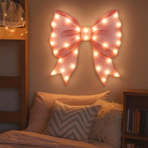 Peut inclure: Une applique murale en forme de nœud rose avec de nombreuses petites ampoules de couleur chaude. La lumière est fixée sur un mur de couleur neutre au-dessus d'un lit avec une literie rose et des coussins décoratifs. Une bibliothèque en bois est visible à droite.