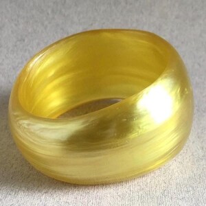 May include: A thick, rounded bangle bracelet in a translucent, golden yellow hue. The bracelet has a smooth, polished surface and a slightly oval shape. The light reflects off the surface, creating a subtle shimmer. This accessory is suitable for everyday wear.