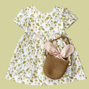 May include: A white dress with a strawberry print and short sleeves. The dress has a gathered skirt and comes with a brown woven basket with pink bunny ears.