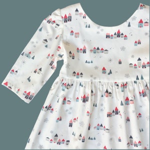 Puede incluir: Un vestido blanco de manga larga y falda fruncida. El vestido tiene un estampado de peque&ntilde;as casas, &aacute;rboles y copos de nieve.