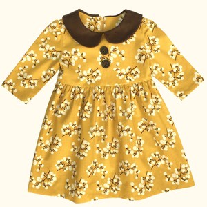 Puede incluir: Un vestido amarillo con estampado floral blanco y cuello de pana marr&oacute;n. El vestido tiene mangas largas y cierre de bot&oacute;n en el cuello.
