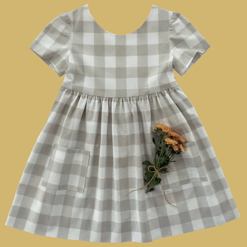 Puede incluir: Un vestido de cuadros vichy blanco y beige con mangas cortas y falda fruncida. El vestido tiene un bolsillo a cada lado y presenta un ramo de flores naranjas.
