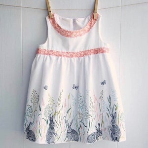 May include: A white sleeveless dress with a pink trim and a floral print of bunnies and butterflies. The dress has a gathered skirt and a round neckline.